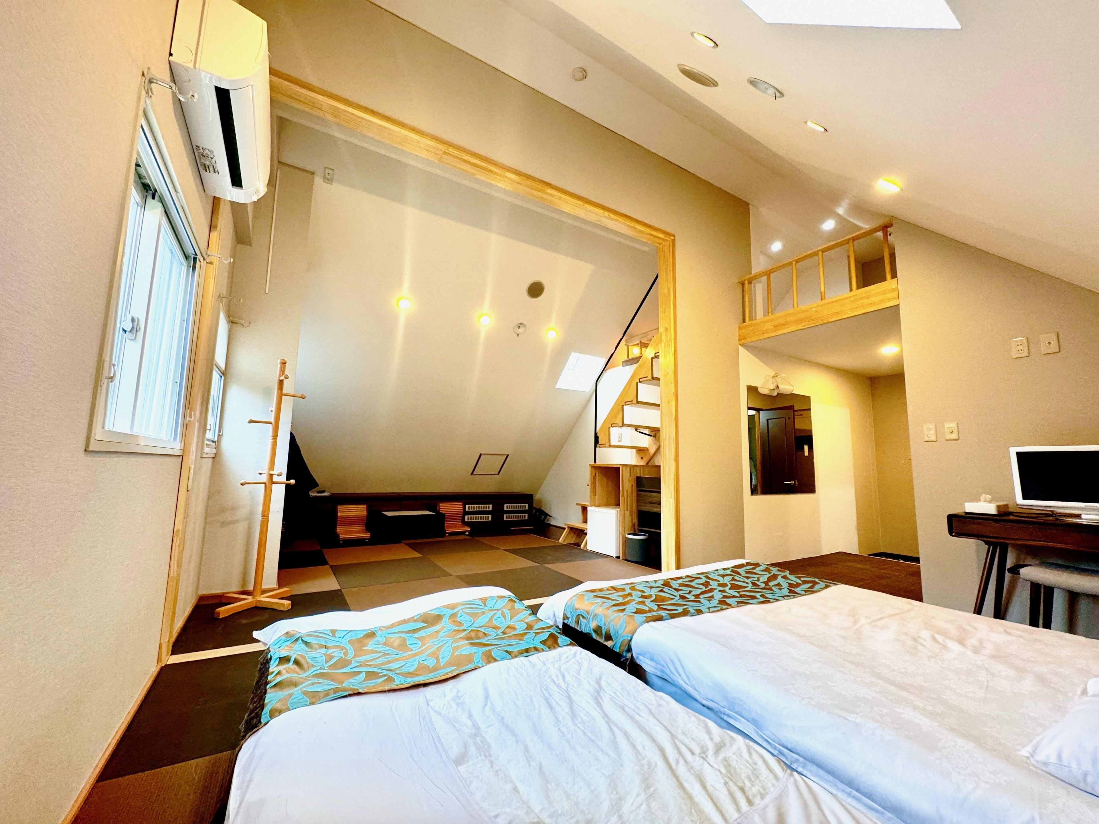 A hotel room with two beds, a desk with a computer, a staircase, and a view of the outside.