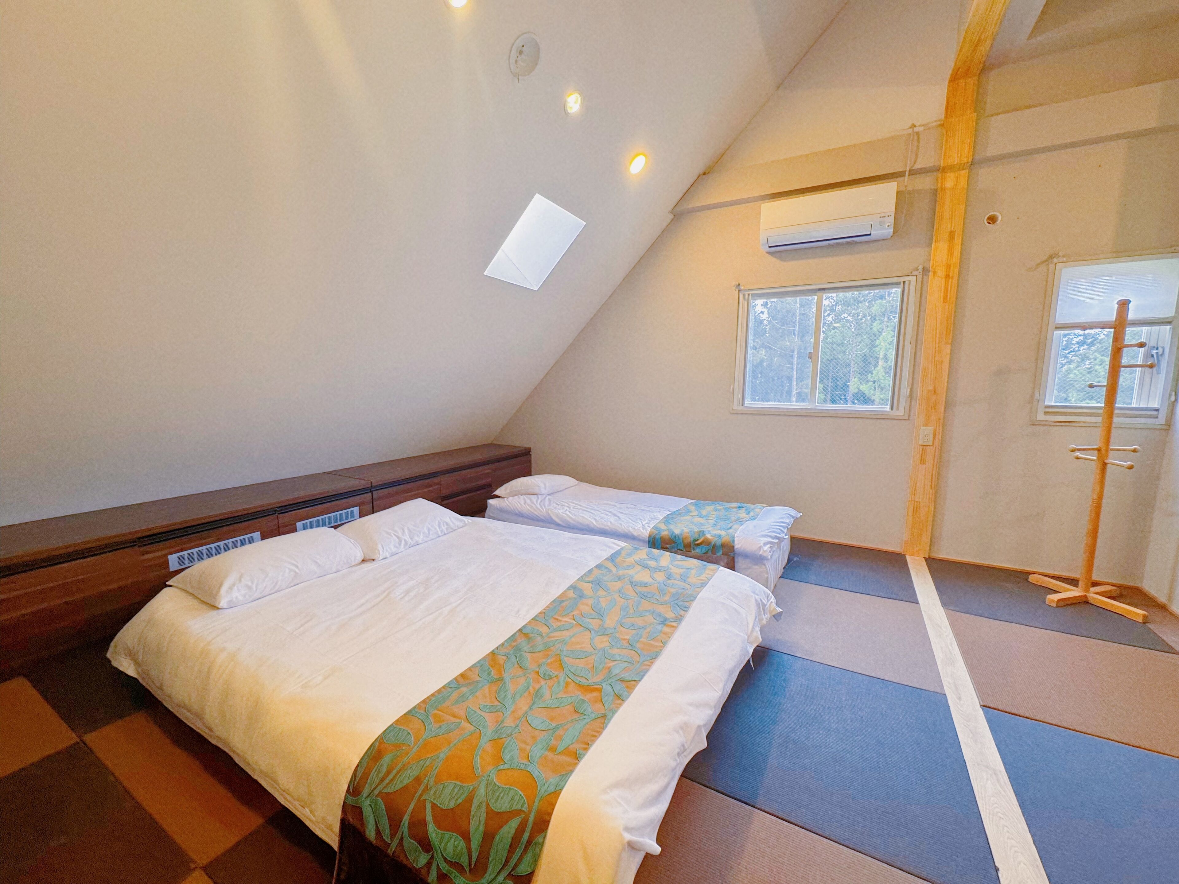 A room with two beds, a wooden headboard, an air conditioning unit, a window, and a wooden rack.