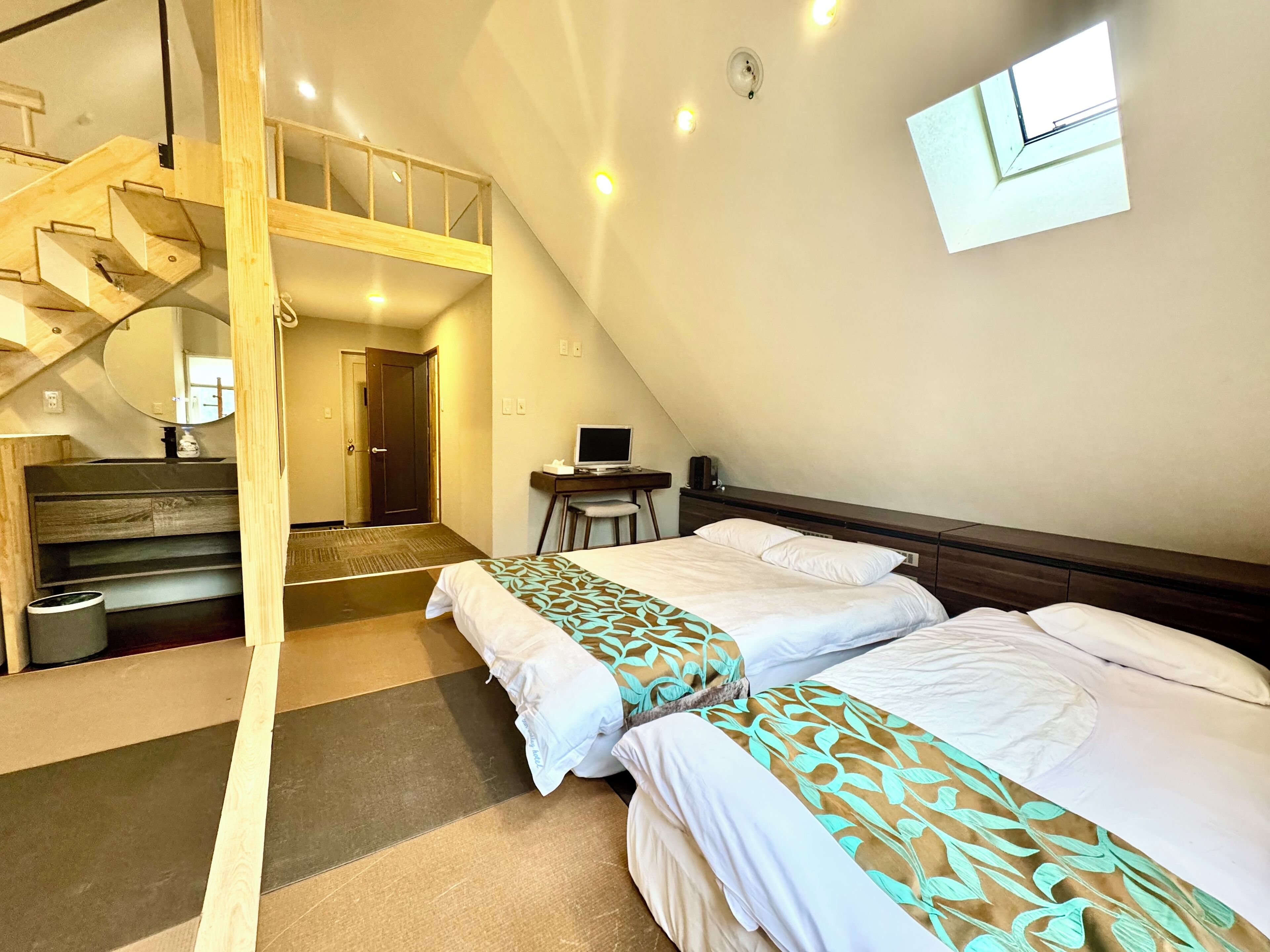 Une pièce avec deux lits, un bureau avec un ordinateur, une petite chaise, un escalier en bois et une porte donnant sur une autre pièce.