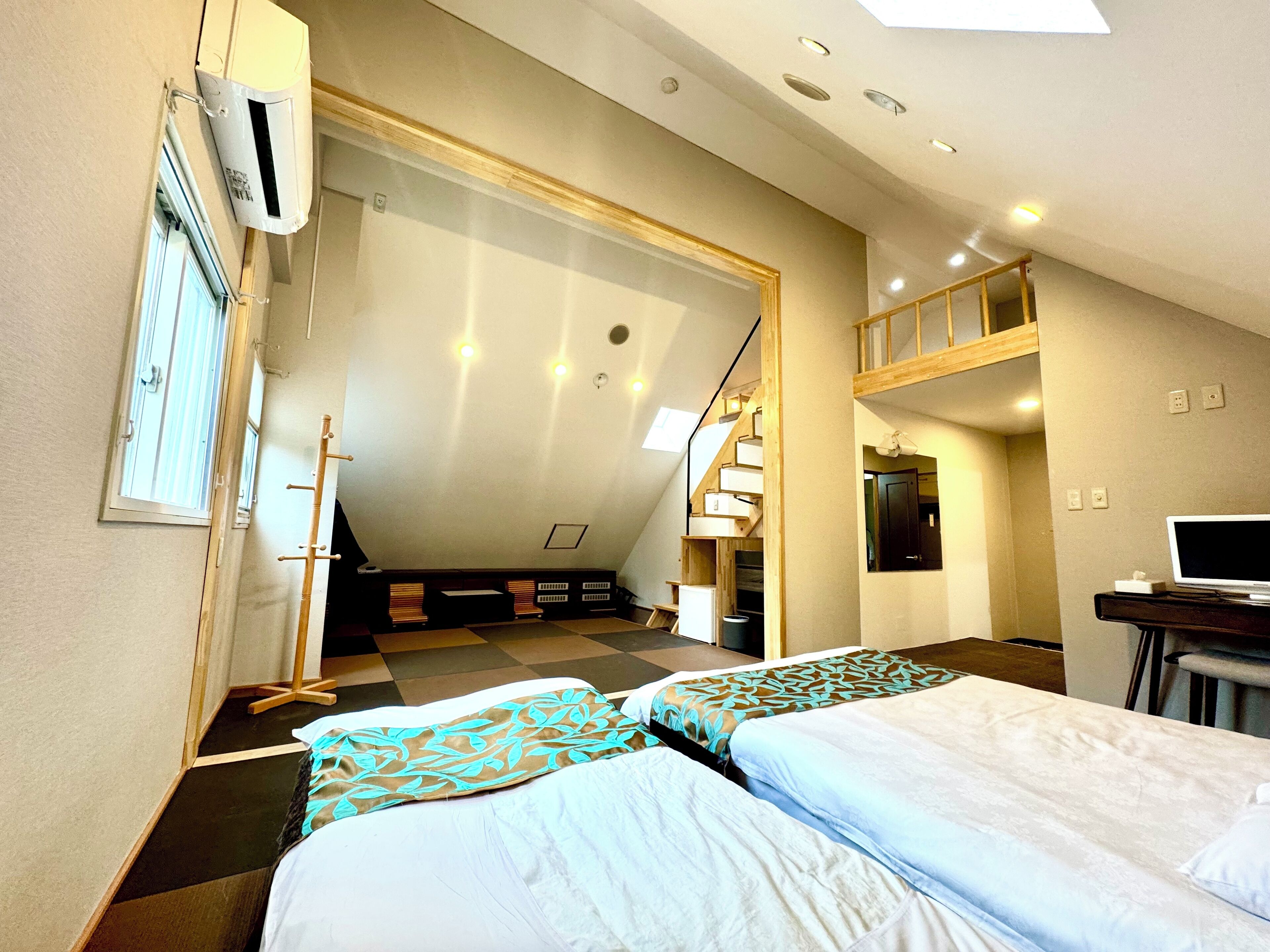 A hotel room with two beds, a desk with a laptop, a staircase, and a large window.