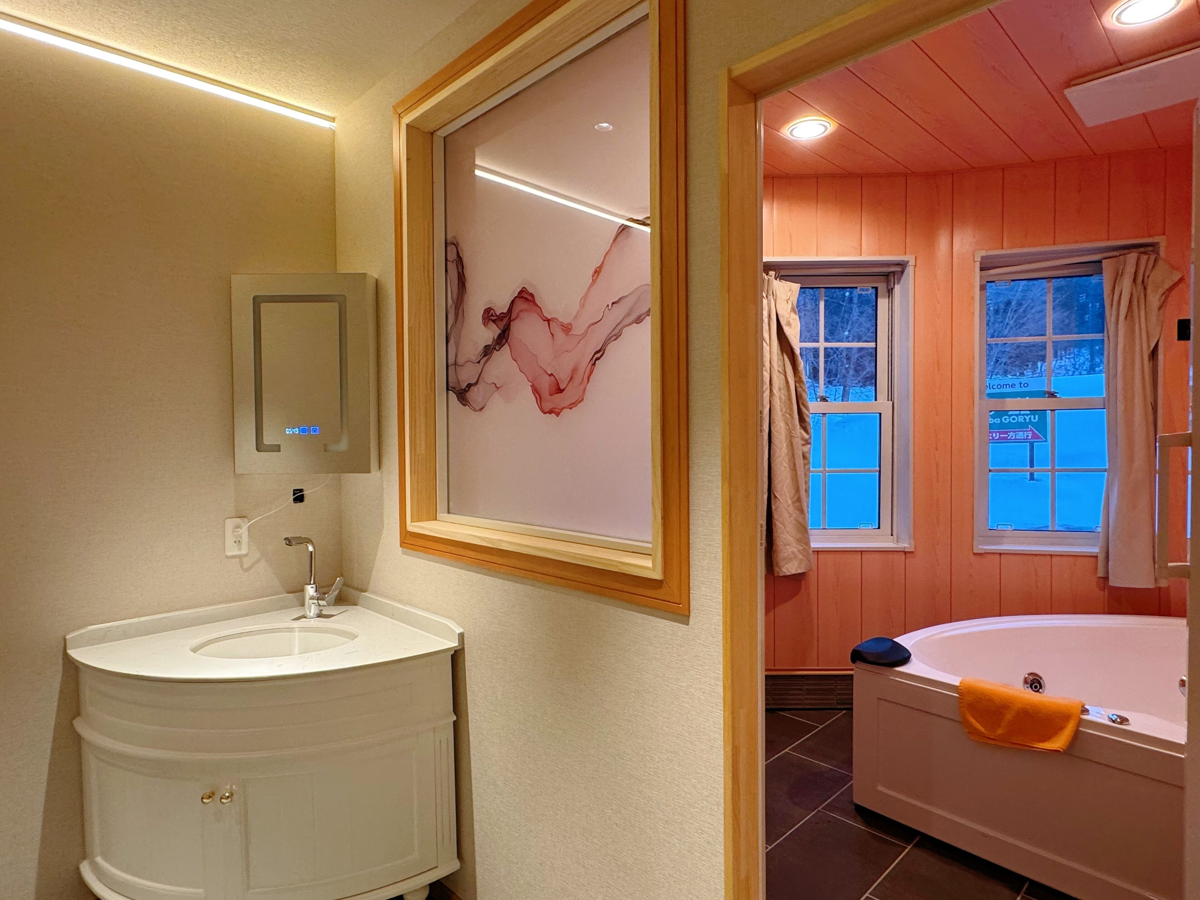 Bagno con mobile lavabo bianco, un'opera d'arte astratta incorniciata e una finestra che si affaccia su un cielo azzurro e alberi.
