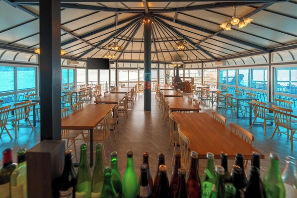 A spacious dining area with multiple wooden tables and chairs, large windows offering views of the sea, and a high ceiling with hanging lights.
