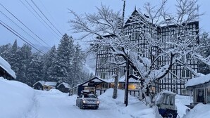 A snow-covered village with traditional timber-framed buildings, a car parked in front, and a small building with a sign.