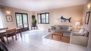 A spacious living area with a sofa, coffee table, and dining table set. There are two windows with open shutters, a potted plant, and a wall-mounted fish decoration.
