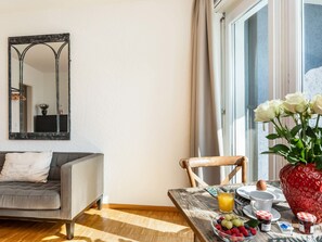 Un salon moderne avec un canapé gris, une table à manger où sont disposés des aliments pour le petit-déjeuner, et une grande fenêtre avec des rideaux.