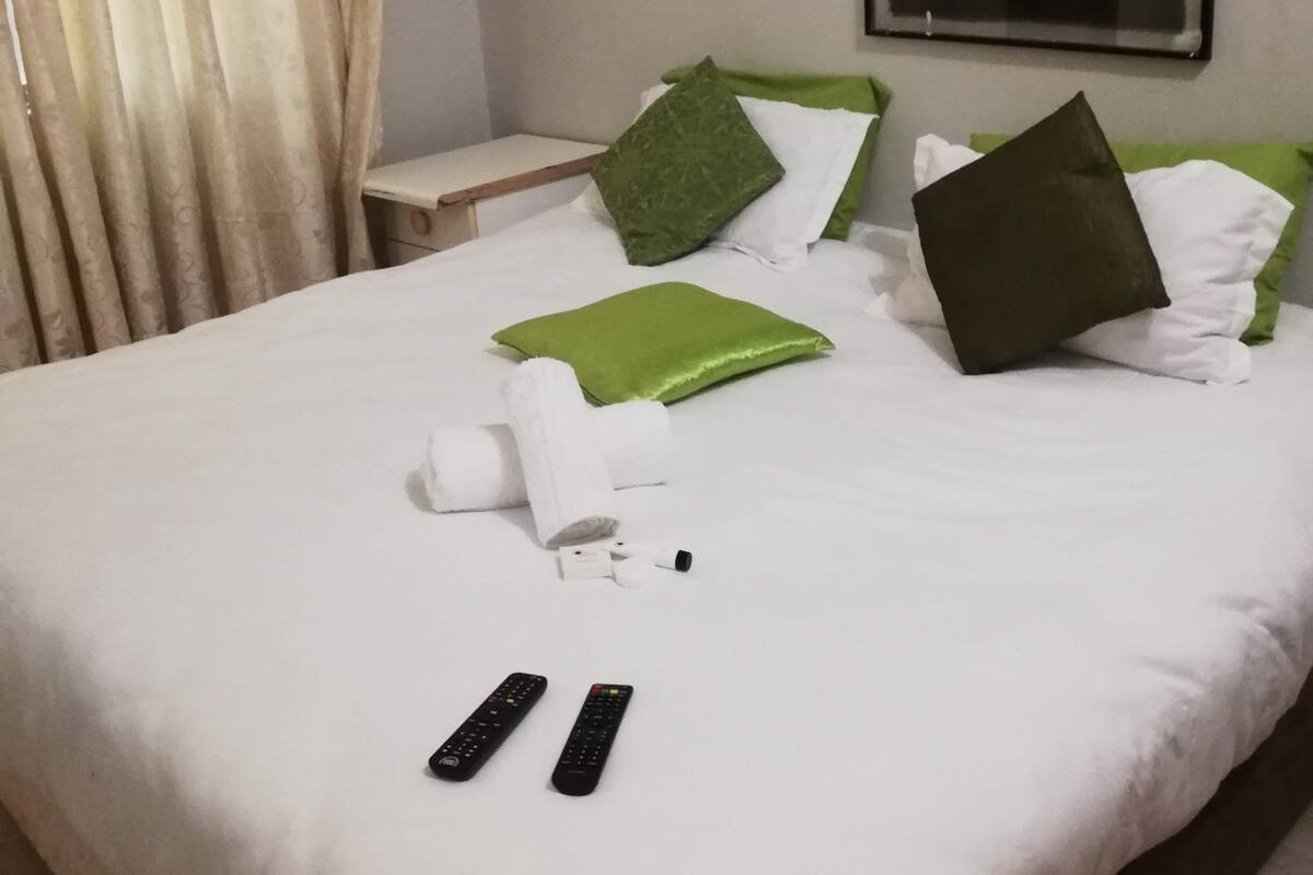 Un lit bien fait, avec une literie blanche, des oreillers verts et blancs, et deux télécommandes posées sur le lit.