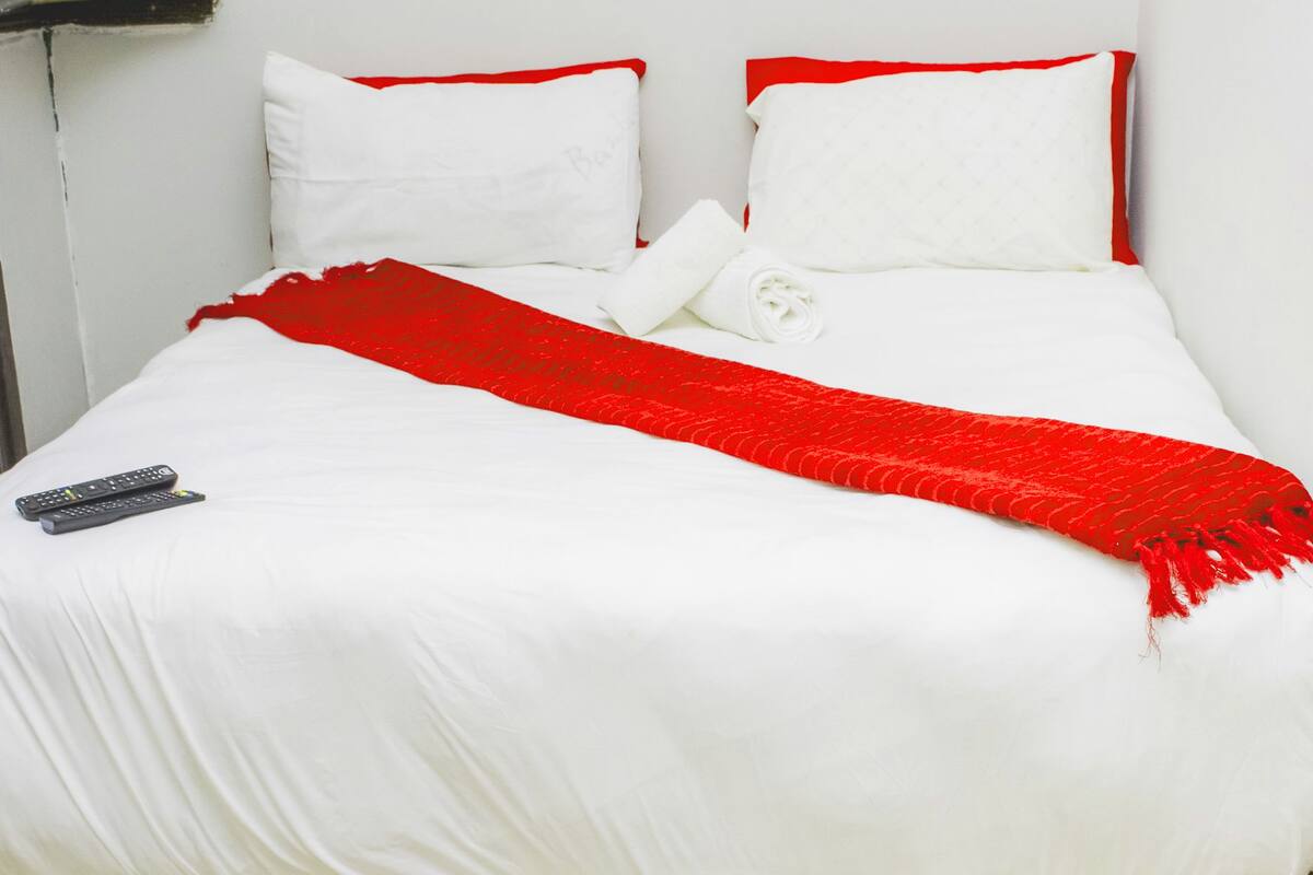 Un lit avec une literie blanche et des touches de rouge, une télécommande posée sur le lit et un plaid rouge.