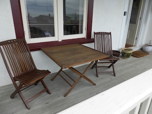 A wooden table and two chairs on a porch with a white railing and a window with a red frame. - Oamaru Holiday Homes 28 Ure Street (Oamaru)