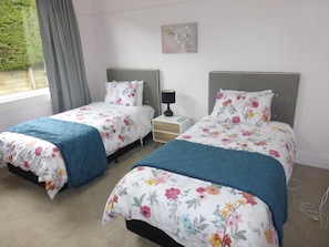 A bedroom with two single beds, each with floral bedding and a blue accent. There is a nightstand with a lamp between the beds, and a window with curtains on the left. - Oamaru Holiday Homes 28 Ure Street (Oamaru)