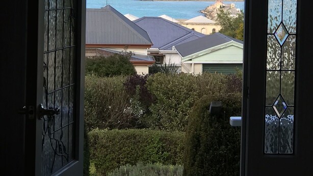 Uitzicht door een deur met een ruitje, waarop een kustlandschap te zien is met huizen, een vuurtoren en de zee.