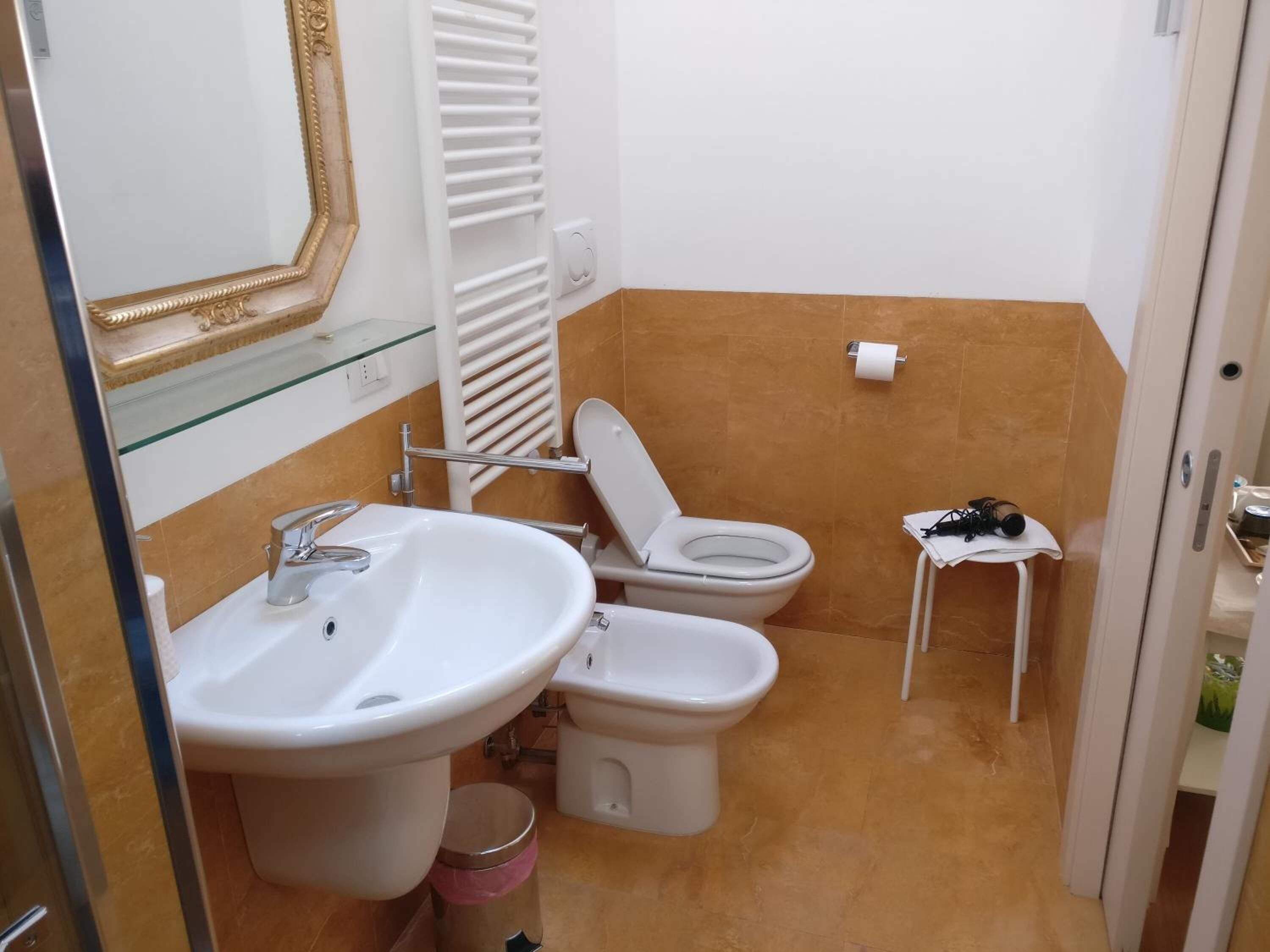Een kleine badkamer met een wastafel, twee toiletten, een spiegel en een tafeltje met een prullenbak.