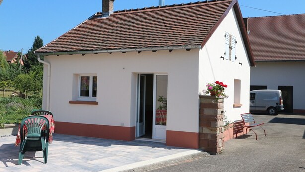 A small, single-story building with a tiled roof, a door, and a small window. There is a chair and a bench outside, and a van parked in the background. - Small studio / Furnished rental (Neuwiller-lès-Saverne)