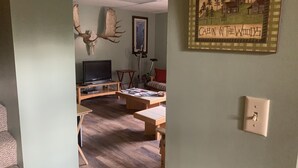 A living room with a television, a couch, a coffee table, and a painting of a cabin in the woods on the wall. - Sawinglogzz Log Cabin (Copper Center)