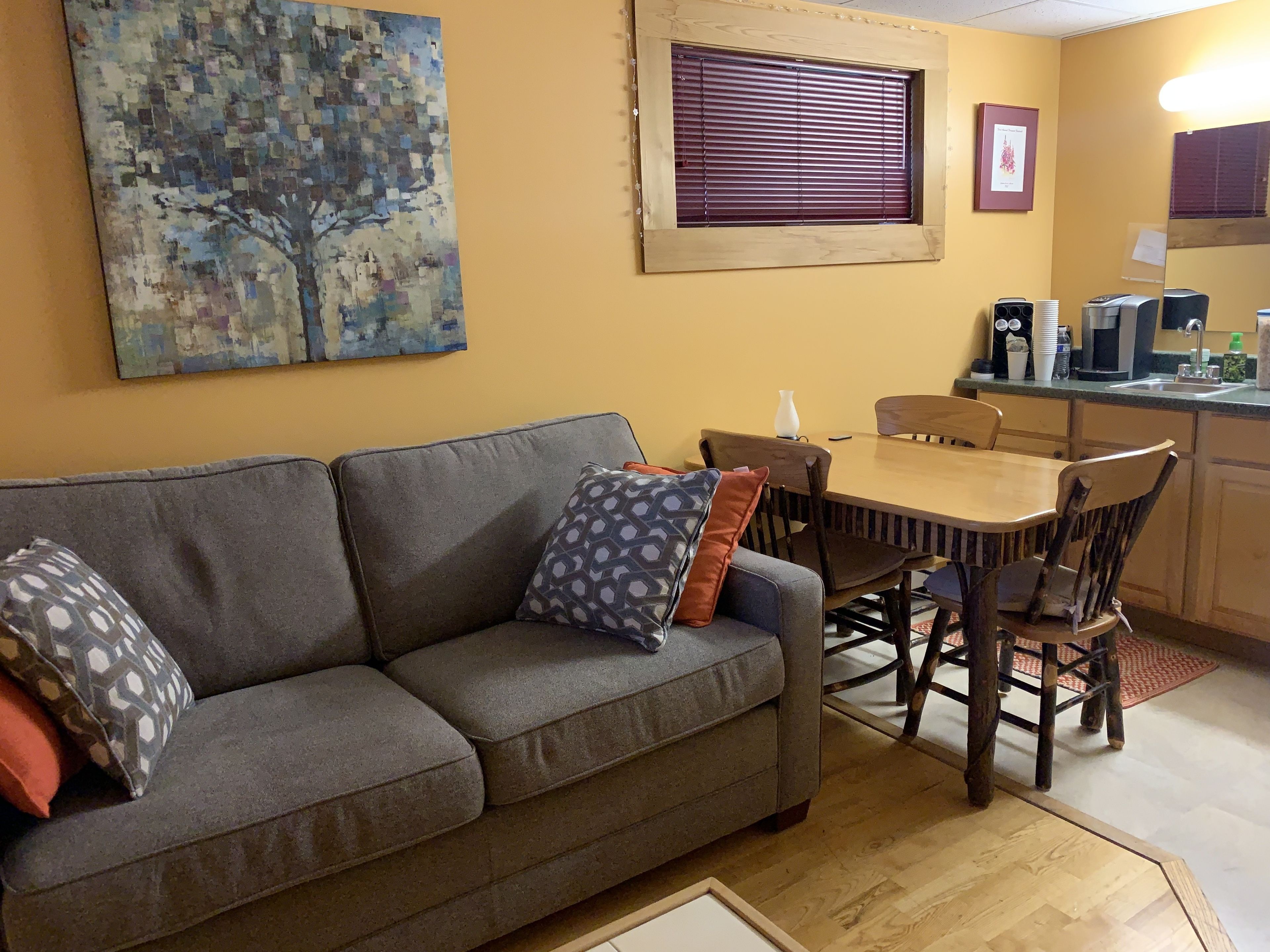 Una sala de estar con un sofá gris, cojines estampados, una mesa de comedor de madera y sillas. Un cuadro de un árbol adorna la pared, y una ventana con persianas se encuentra sobre la mesa.