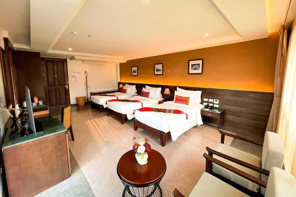 Quarto de hotel com duas camas, uma escrivaninha, uma cadeira, uma mesinha de centro com um vaso, e cabeceira de madeira.