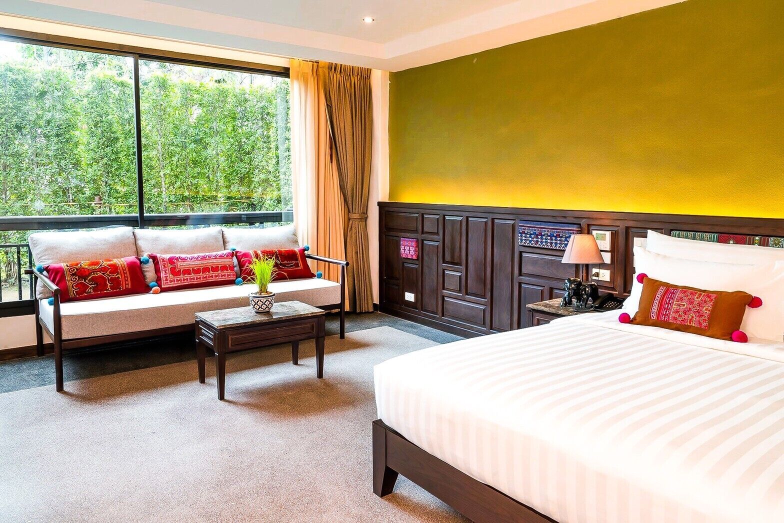 Una habitación de hotel con cama, un sofá con cojines de colores, una mesita con una planta y vistas a zonas verdes por la ventana.