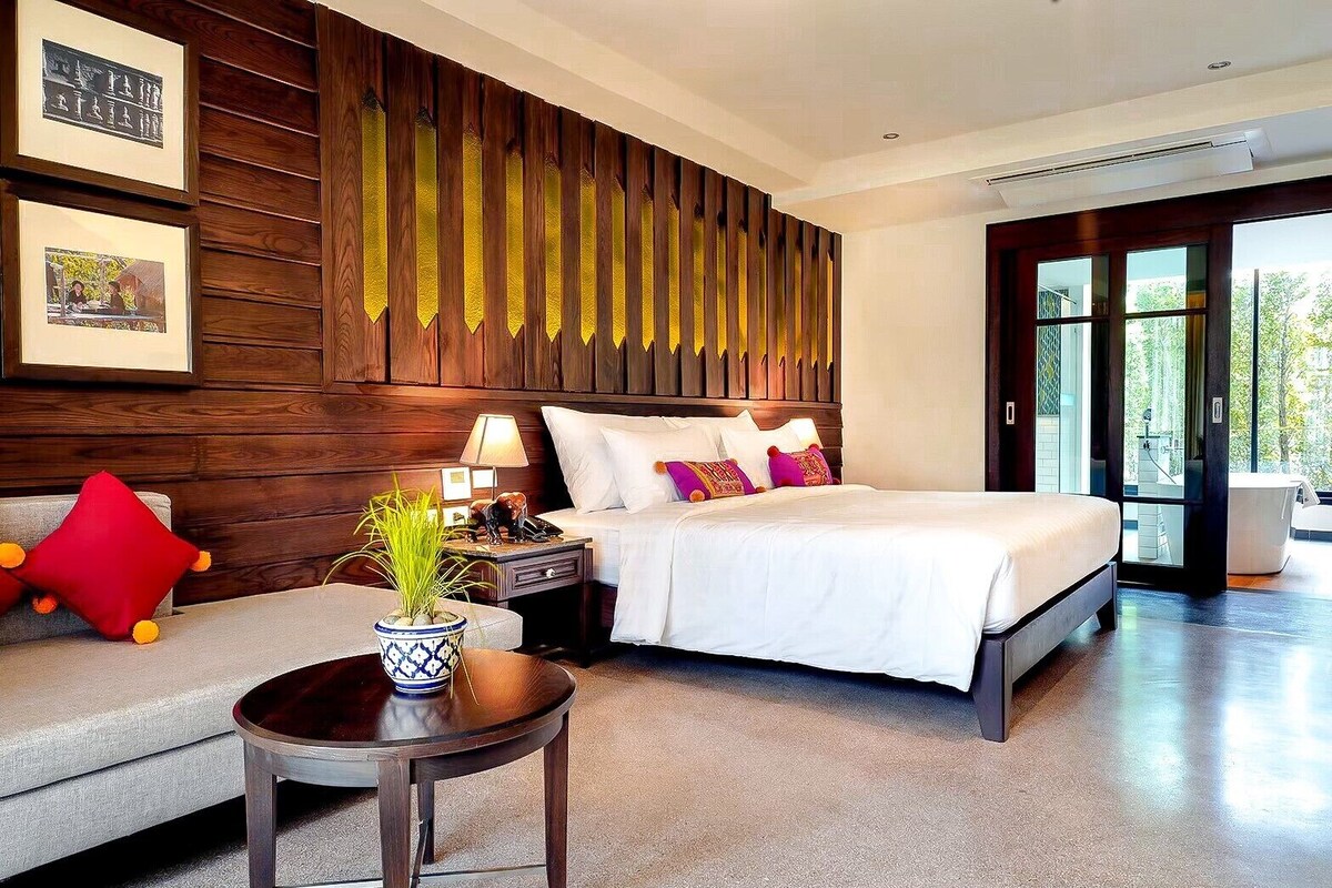 Quarto de hotel moderno com uma cama grande, um sofá, mesa de cabeceira e planta em vaso.