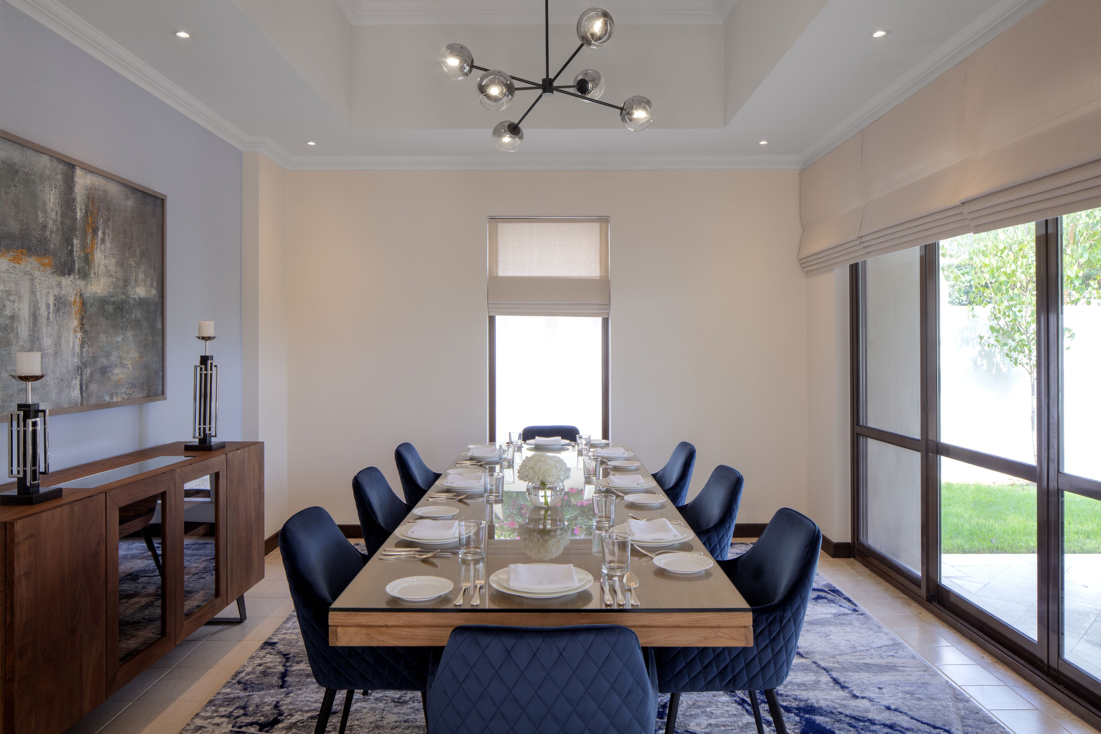 A dining room with a long table set for six, surrounded by six chairs, a chandelier, and large windows.