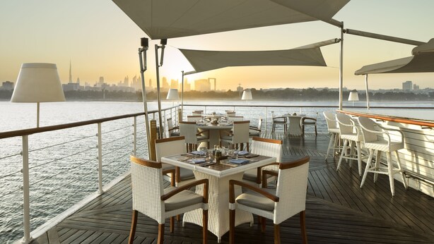 Un espace repas sur un bateau, avec des tables et des chaises, une rambarde et une vue sur les toits d’une ville qui se dessinent à l’horizon, de l’autre côté de l ’eau.