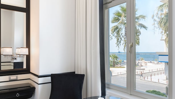 Habitación de hotel con un ventanal arqueado grande, una silla negra, una mesa redonda con dos tazas blancas y suelo de madera.