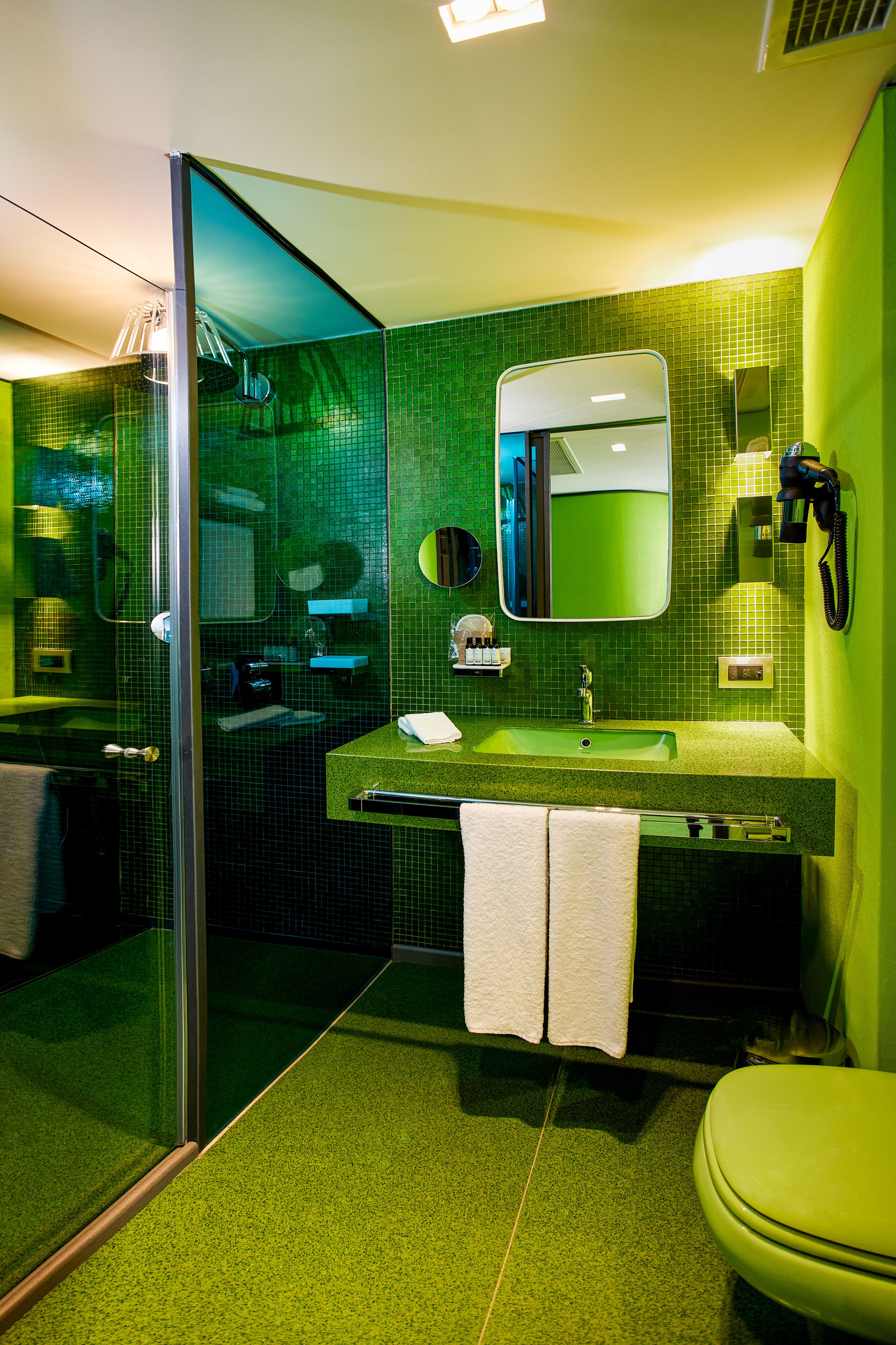 Um banheiro com parede de azulejos verdes, pia com espelho acima, vaso sanitário e box de vidro.