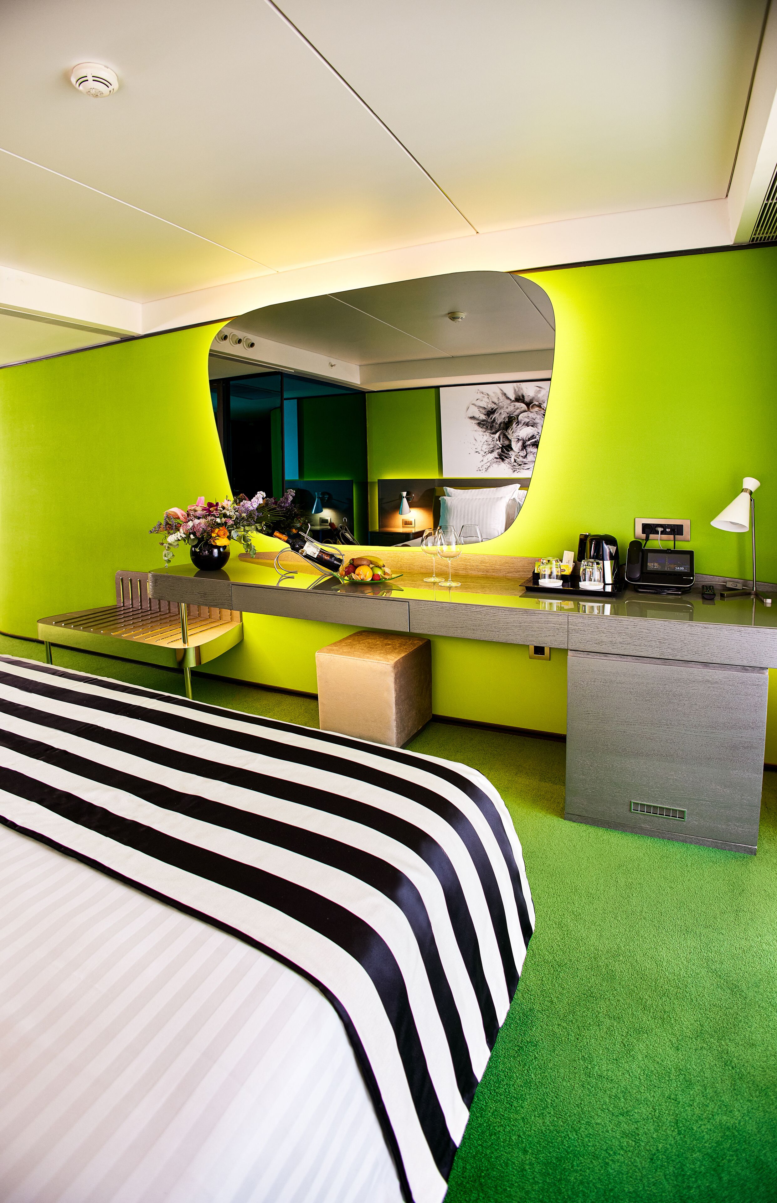 A modern hotel room with a green wall, a bed with striped bedding, a desk with a mirror, a coffee maker, and a vase with flowers.
