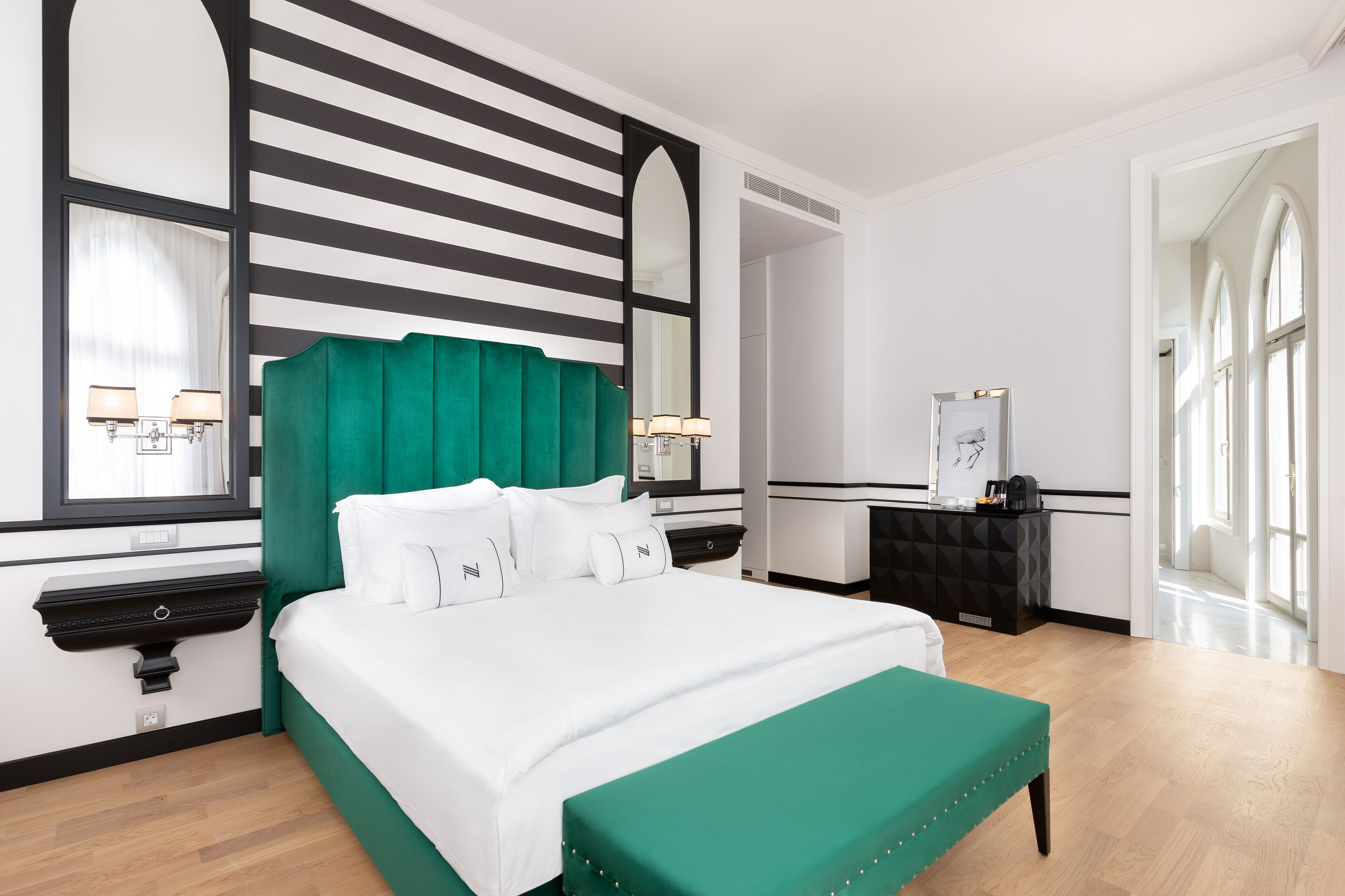 A hotel room with a large bed, a green upholstered bench, a black and white striped wall, and a large mirror.