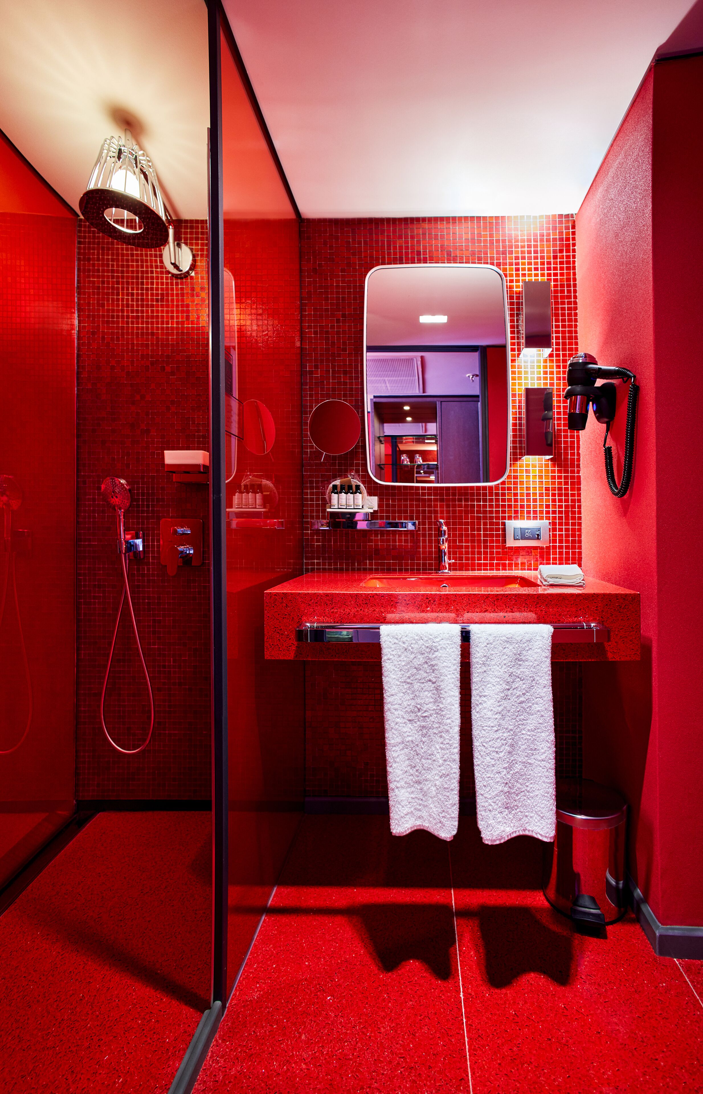 Un baño moderno con pared de azulejos rojos, cabina de ducha de vidrio, lavabo con espejo y secador de pelo montado en la pared.
