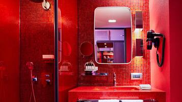Un baño moderno con pared de azulejos rojos, cabina de ducha de vidrio, lavabo con espejo y secador de pelo montado en la pared.