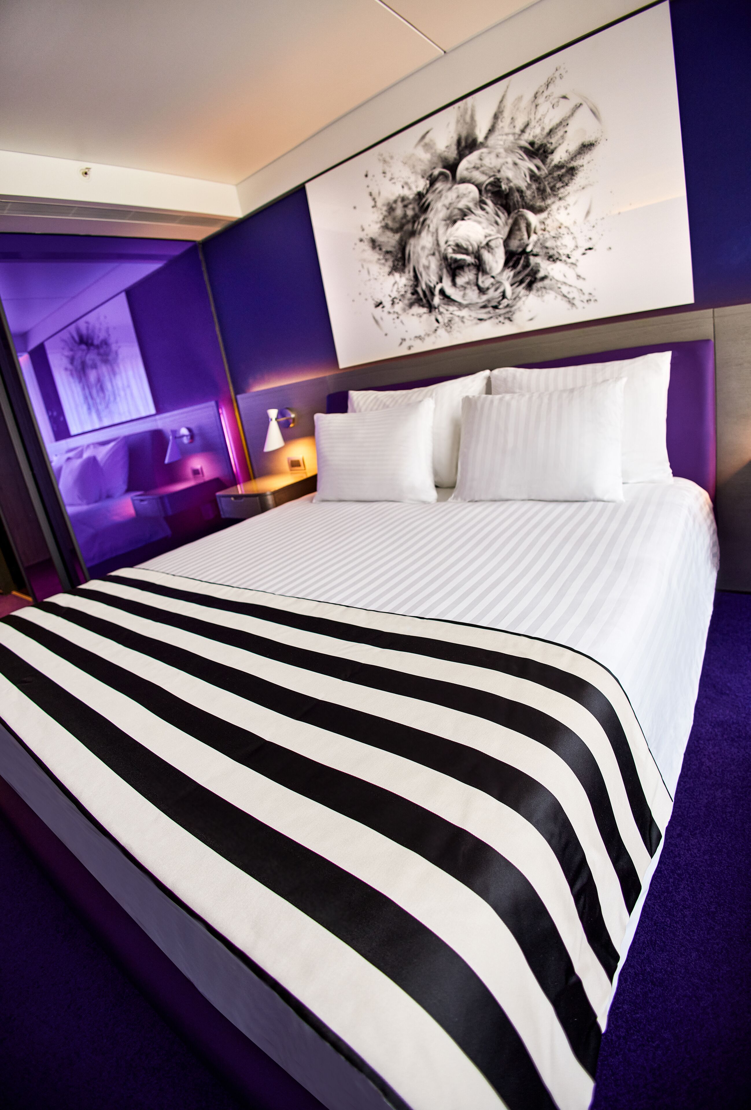 Habitación de hotel con una cama grande, cabecera con una obra de arte con la figura de un león e iluminación ambiental morada.