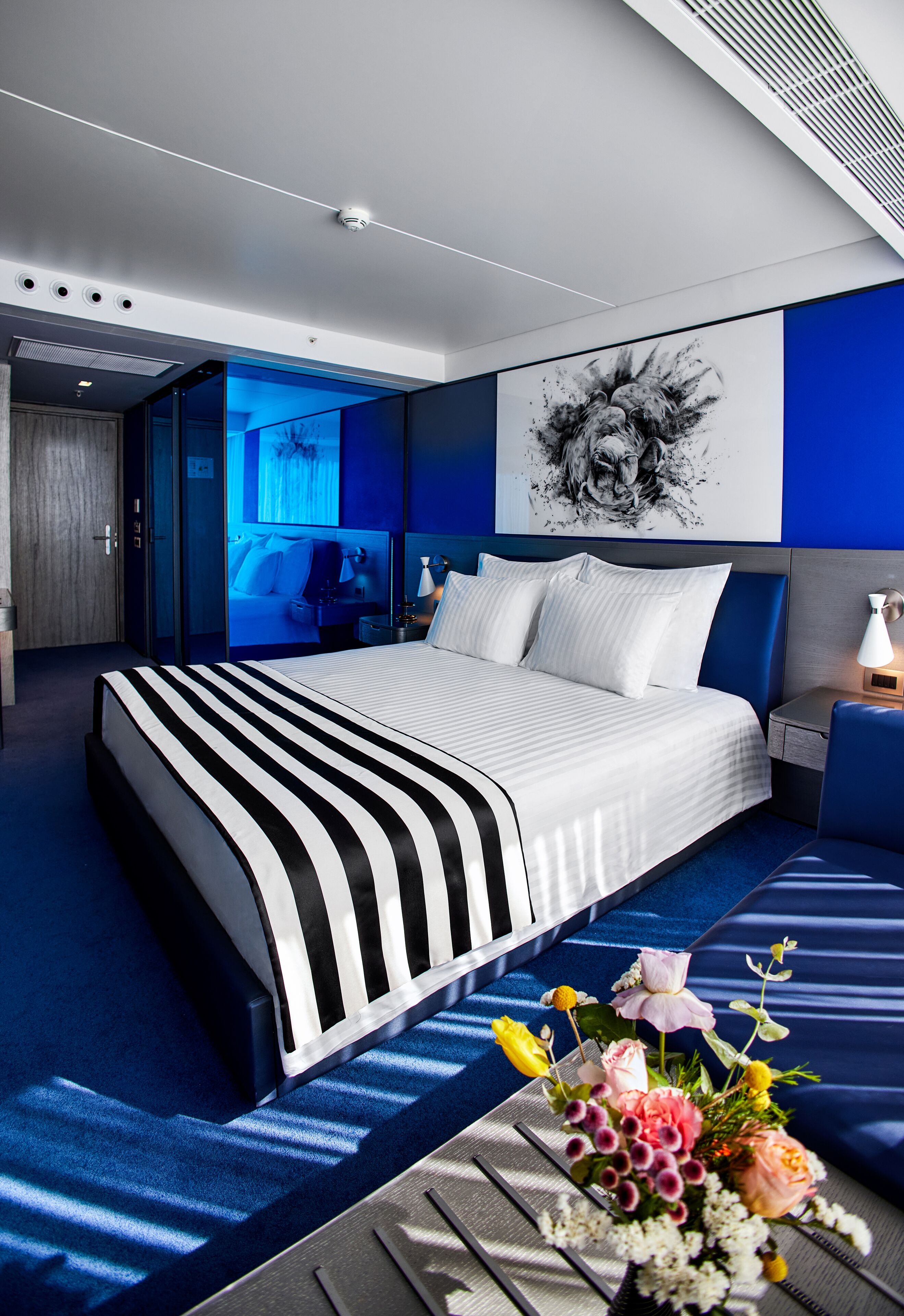 A modern hotel room with a large bed, a nightstand, a desk, and a bathroom visible through a glass door.