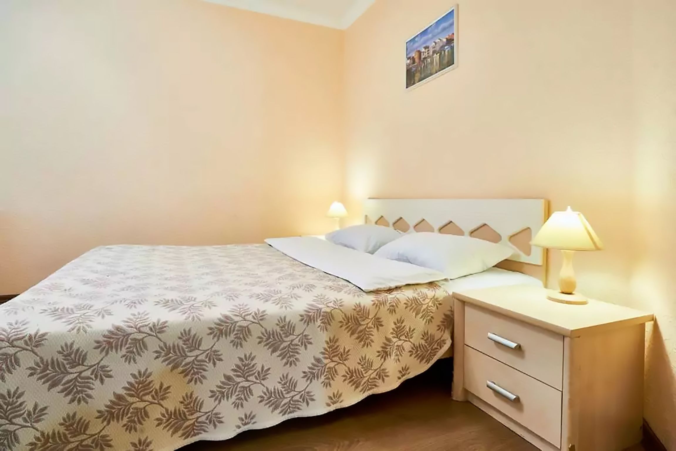 A neatly made bed with a patterned bedspread, a bedside table with a lamp, and a small drawer.