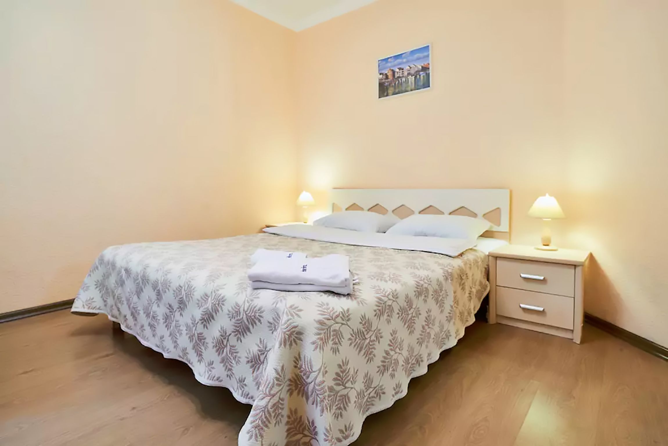 A neatly made bed with a floral bedspread, two bedside lamps, and a small bedside table with drawers.
