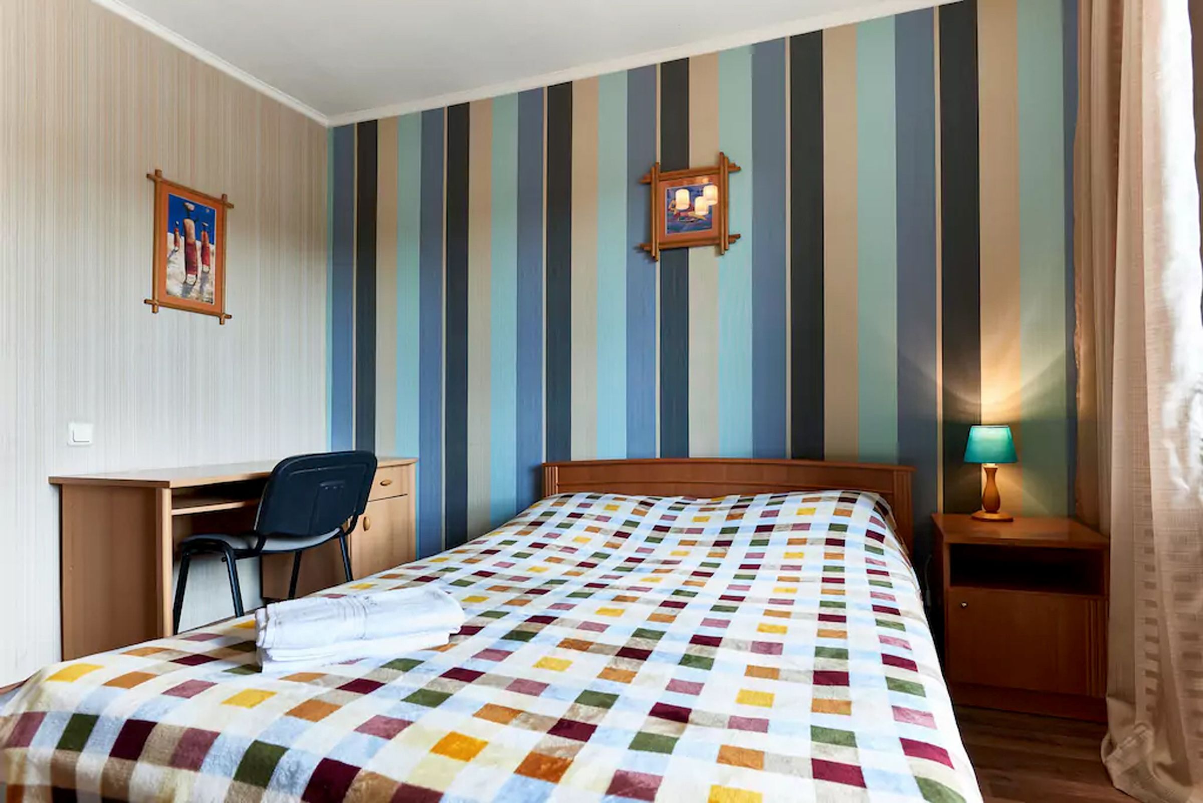A hotel room with a bed, a desk, a chair, and a nightstand with a lamp.