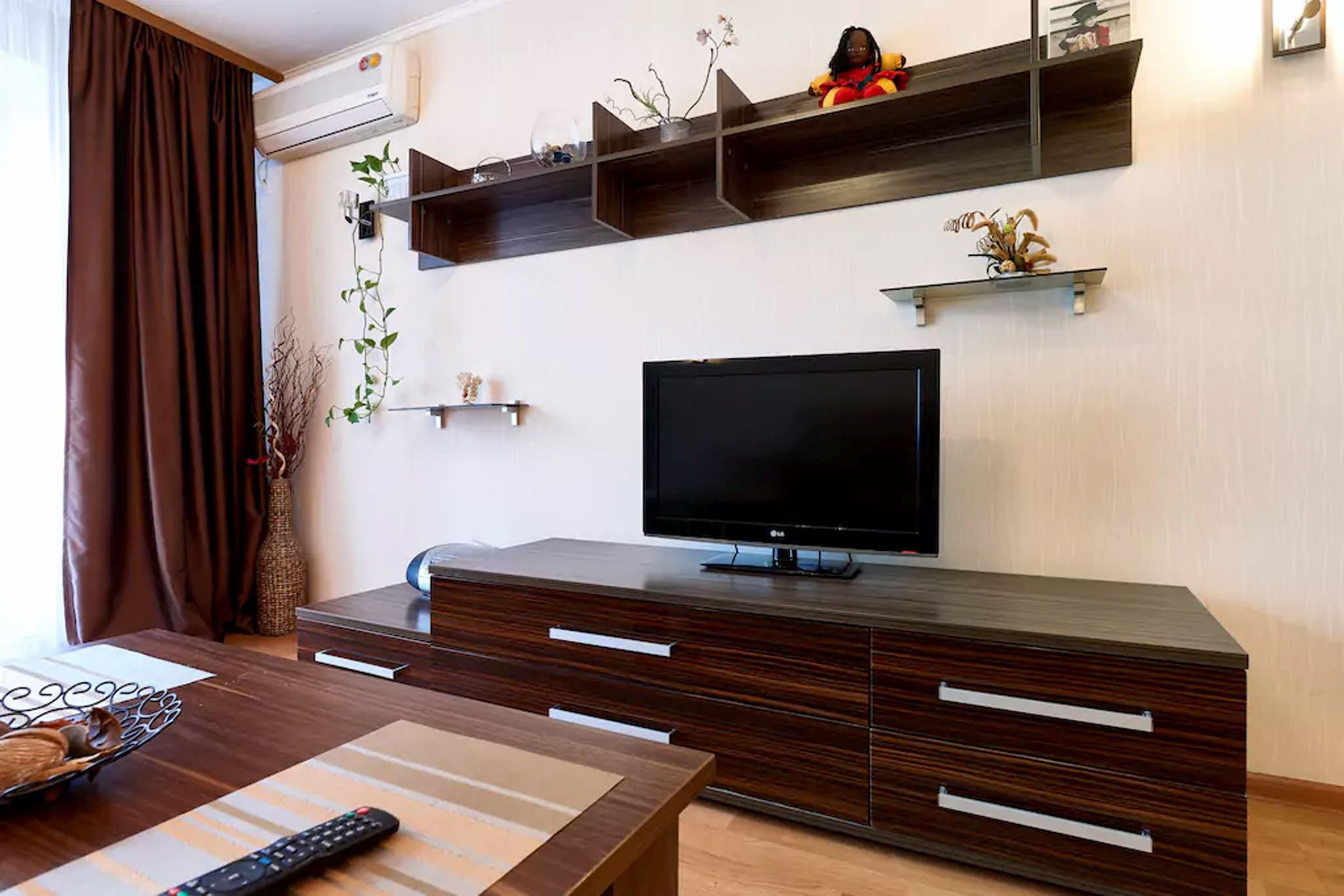 A modern living room with a flat-screen TV, wooden entertainment unit, and a dining area with a table and chairs.