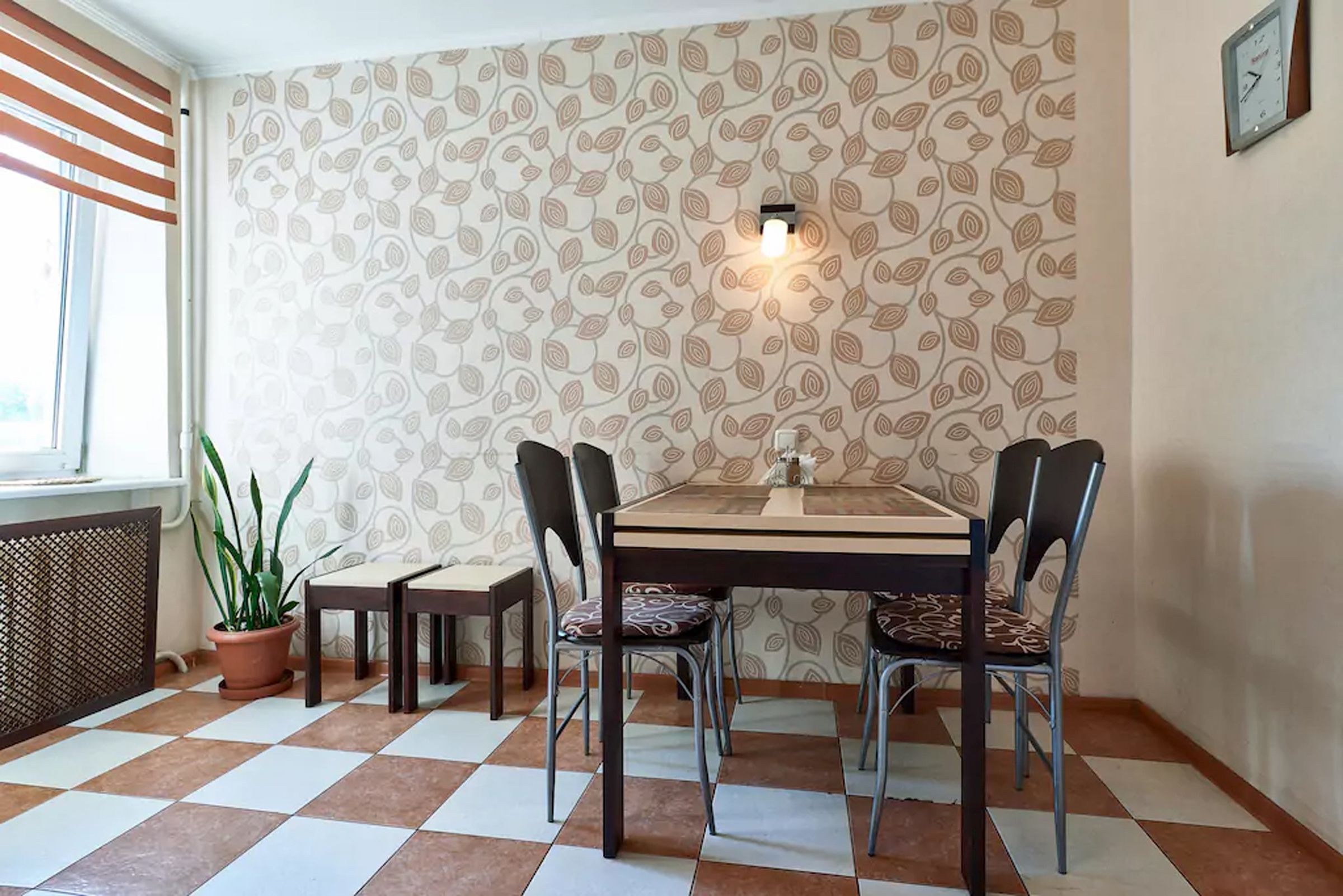 A dining area with a checkered floor, patterned wallpaper, a table with chairs, a potted plant, and a wall-mounted light fixture.