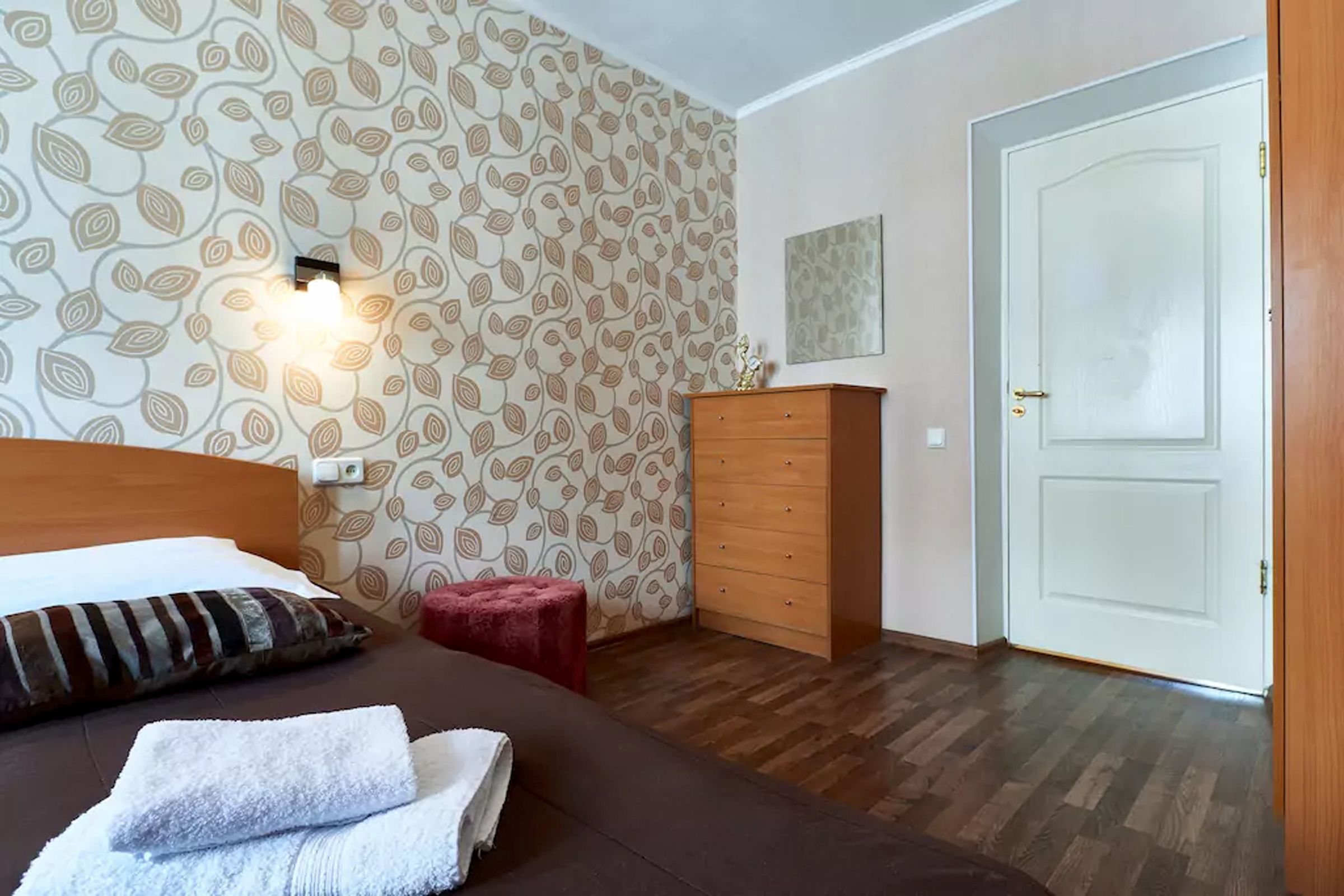 Habitación de hotel con papel tapiz estampado, cómoda de madera, taburete rojo, cama con almohada a rayas y una puerta blanca.