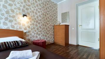 Habitación de hotel con papel tapiz estampado, cómoda de madera, taburete rojo, cama con almohada a rayas y una puerta blanca.
