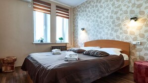 A neatly made bed with a brown bedspread and pillows, a bedside table with a lamp, and a window with blinds. - Home-Hotel Nizhniy Val 41-2 (Kyiv)