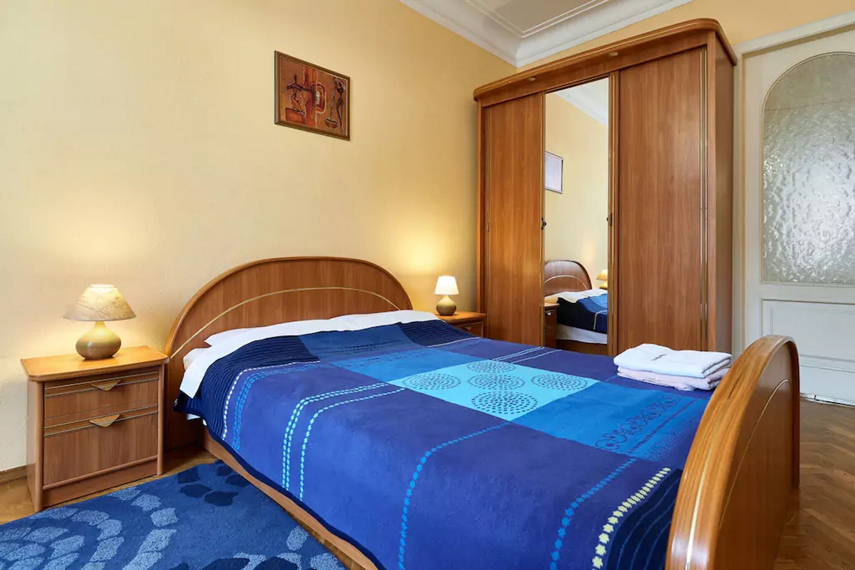 Una cama bien hecha con colcha azul y blanca, una mesita de noche de madera con una lámpara y un armario de madera con espejo.