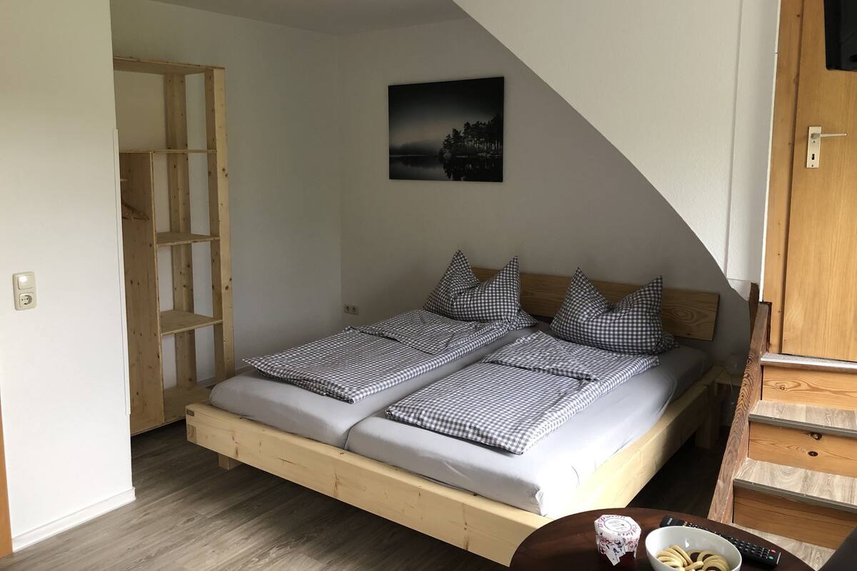 Une chambre avec un lit en bois, une literie à carreaux, une table de chevet avec un bol et une télécommande, et un tableau accroché au mur.