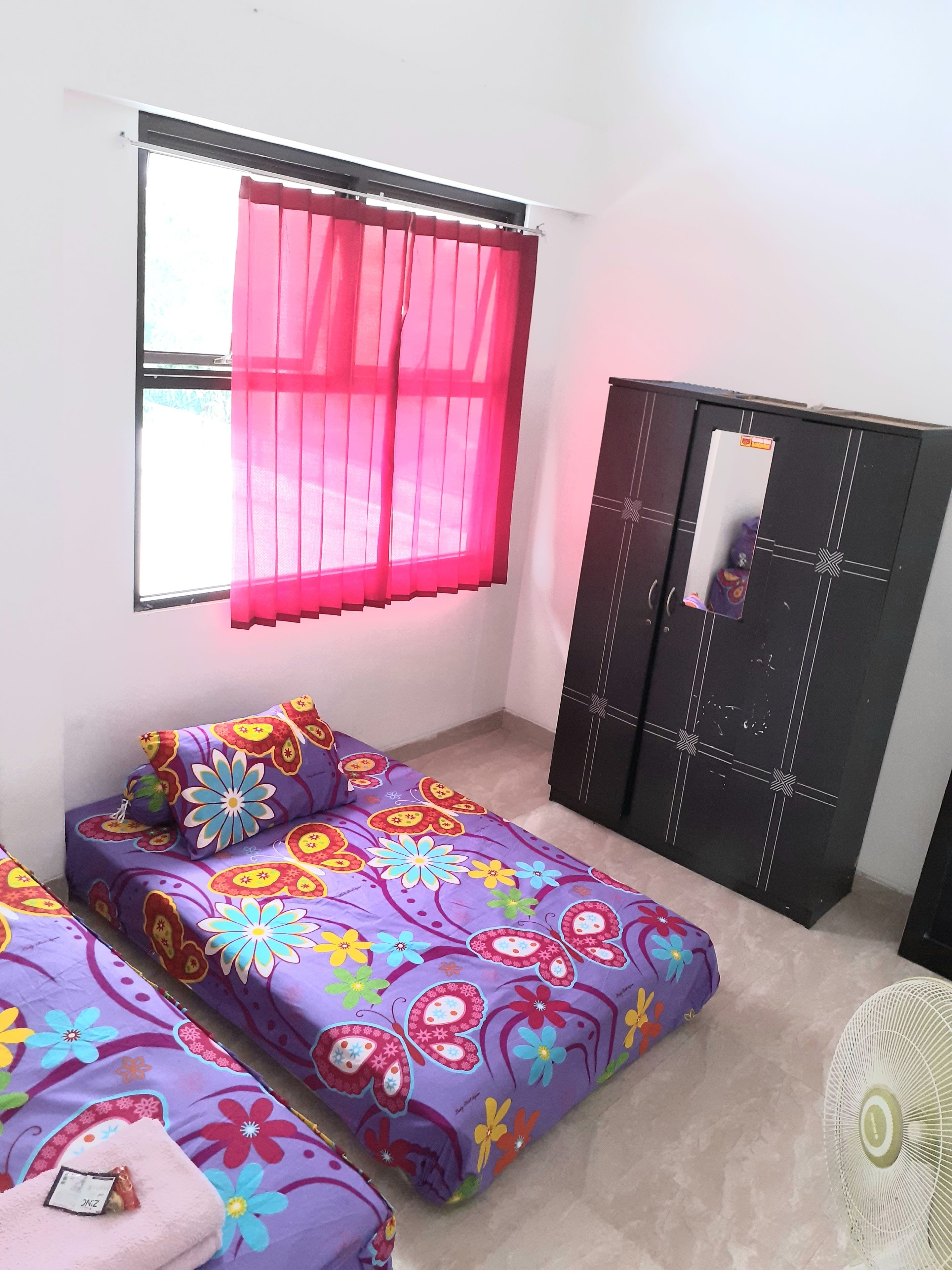 Ein Zimmer mit zwei Betten, einem Fenster mit rosa Vorhängen, einem schwarzen Kleiderschrank und einem weißen Ventilator.