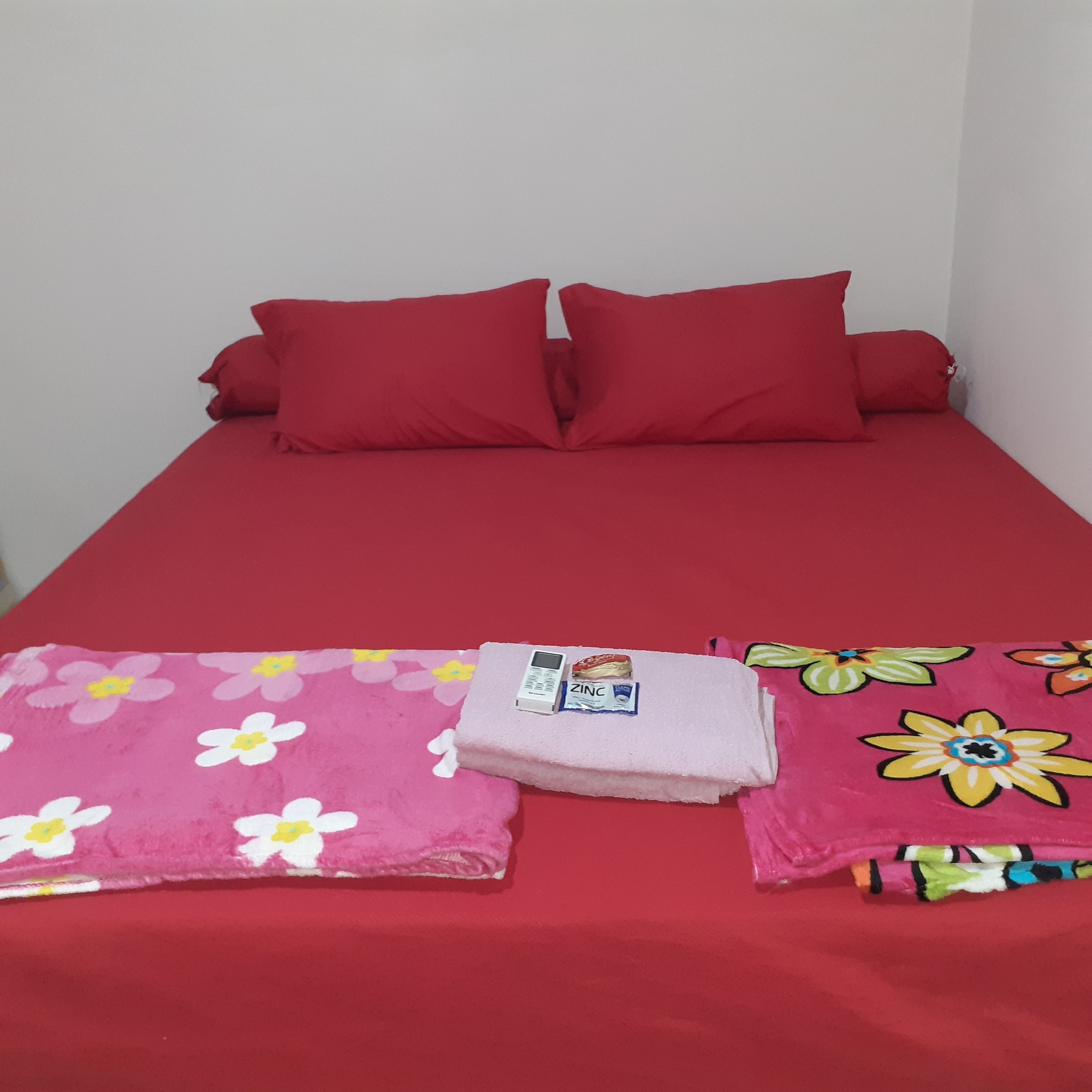 A bed with red bedding, a pink floral blanket, and a pink towel.