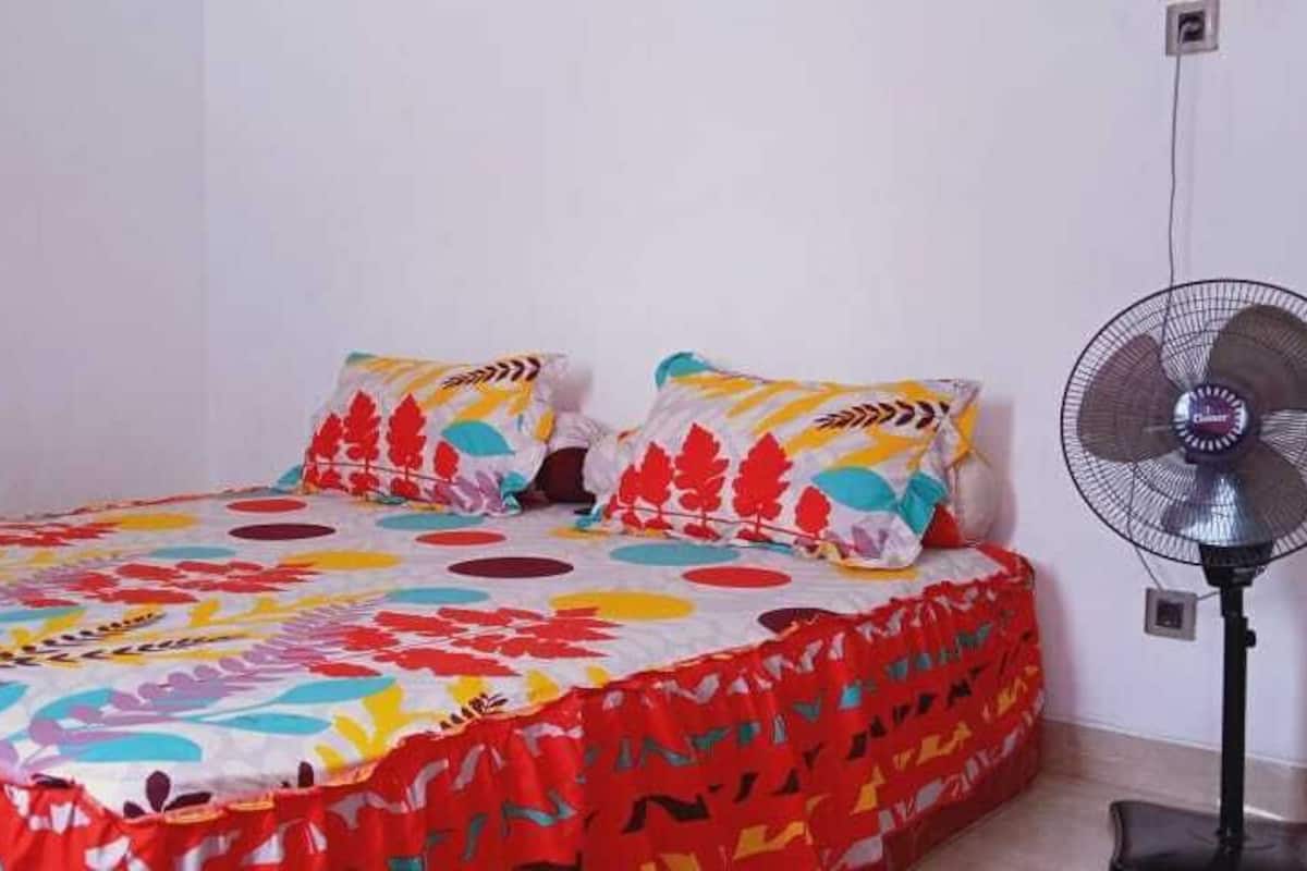 A bed with a colorful bedspread and two pillows, a standing fan, and a power outlet on the wall.