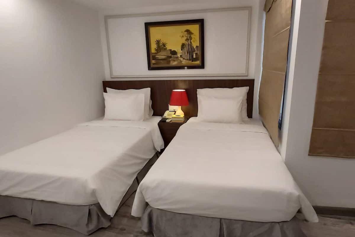 A hotel room with two single beds, a nightstand with a lamp, a framed picture on the wall, and a window with a blind.