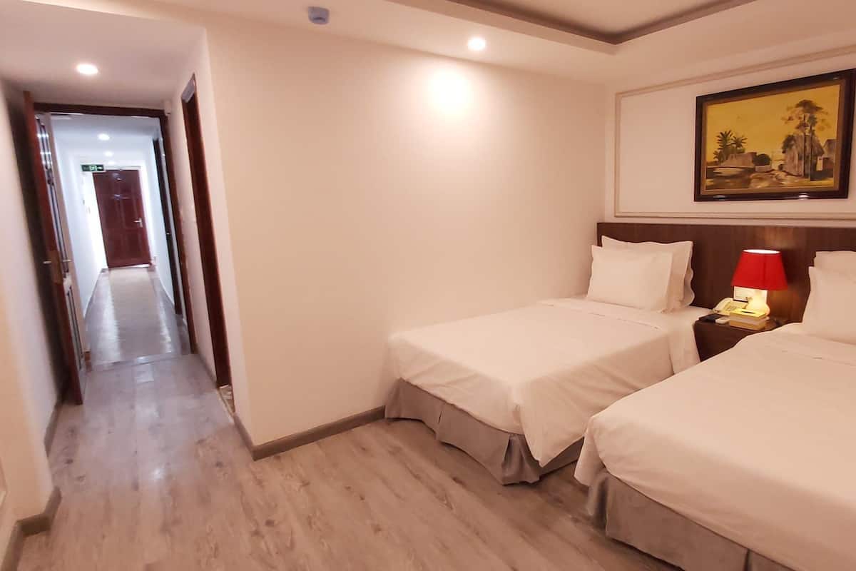 A hotel room with two single beds, a nightstand with a lamp, a painting on the wall, and a hallway leading to another room.