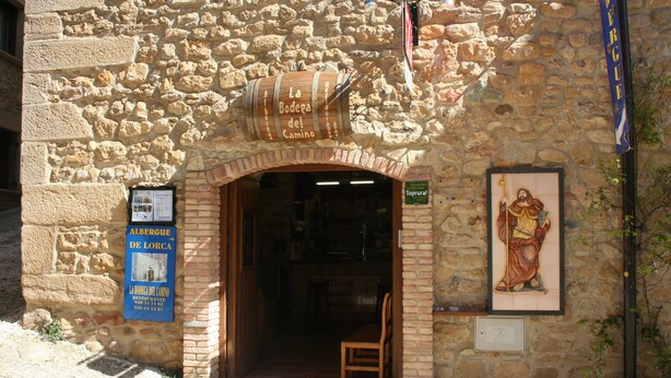 A stone building with a wooden barrel sign reading "La Bodega del Camino" and a framed religious figure on the wall.