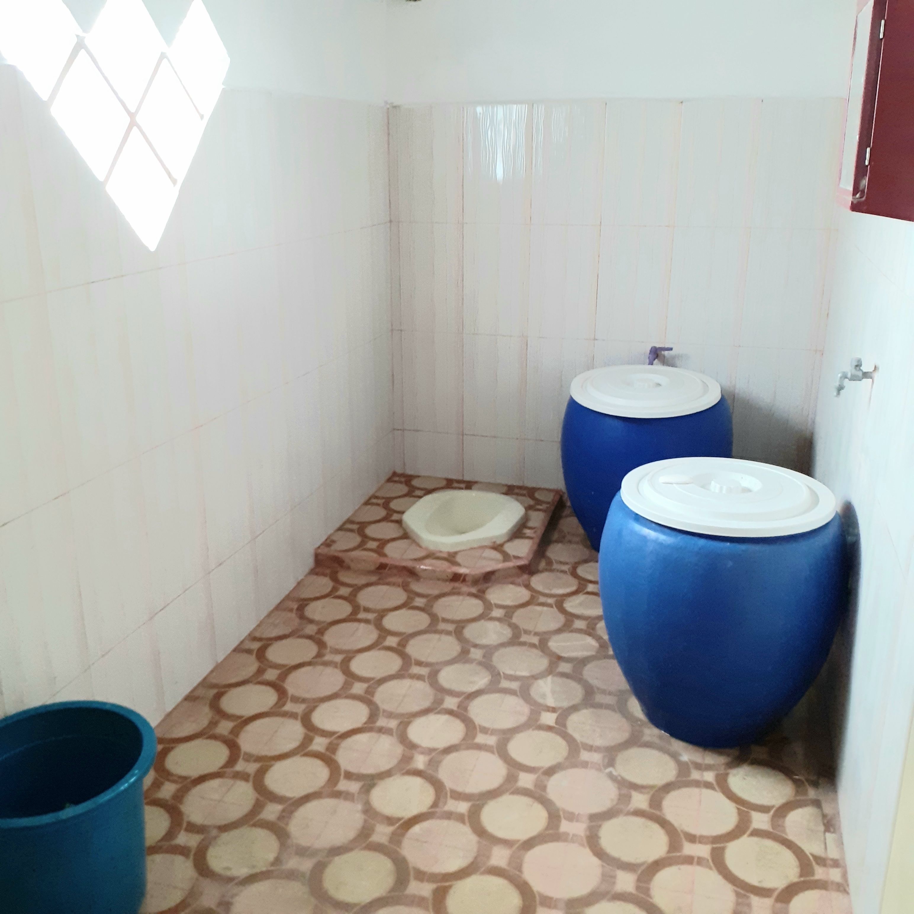 A small, tiled bathroom with a toilet, a blue water tank, and a blue bucket.