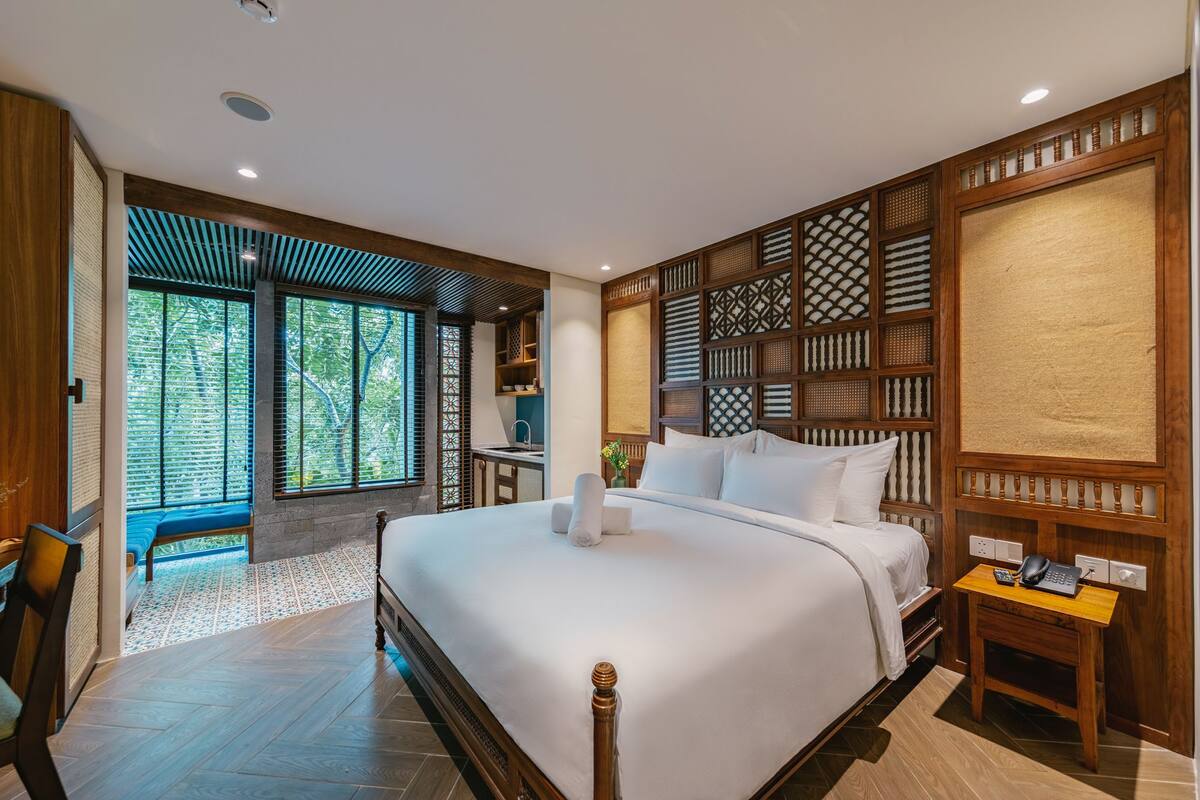 A spacious bedroom with a large bed, a wooden headboard, a small desk with a telephone, and a view of the outdoors through a large window.