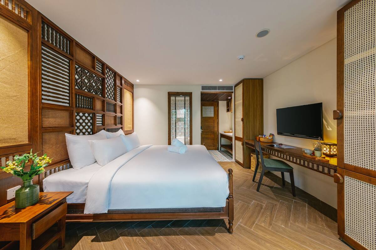 A hotel room with a large bed, a desk with a chair, a television, and a decorative wooden partition.